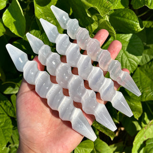 Hand holding multiple spiral selenite wands with pointed tips, glowing under natural sunlight.