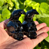 Polished black obsidian carvings featuring a raven perched on a human skull, displayed on a hand.  