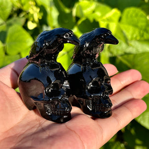 Polished black obsidian carvings featuring a raven perched on a human skull, displayed on a hand.  